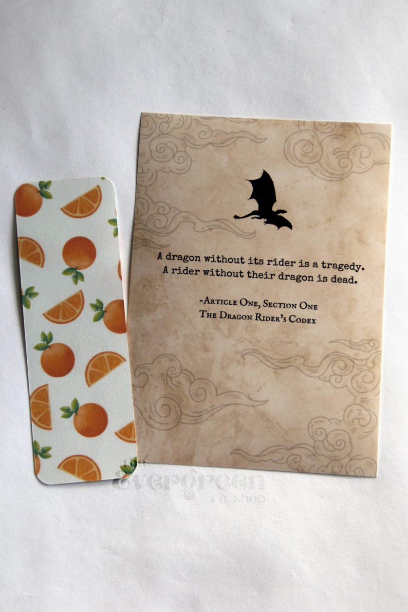 May include: A bookmark with orange slices and a card with a dragon silhouette and text. The bookmark features a white background with orange slices and green leaves. The card has a dragon silhouette and text that reads "A dragon without its rider is a tragedy."