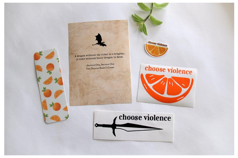 May include: A collection of stickers and a bookmark with orange and dragon-themed designs. The bookmark features oranges, while the stickers display an orange slice and the phrase "choose violence" with a sword graphic. A card with a dragon silhouette and quote is also present.