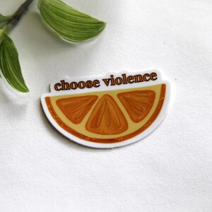 May include: A sticker featuring a slice of orange with the words "choose violence" in brown text. The orange slice is orange and brown with a white border. Green leaves are in the background.