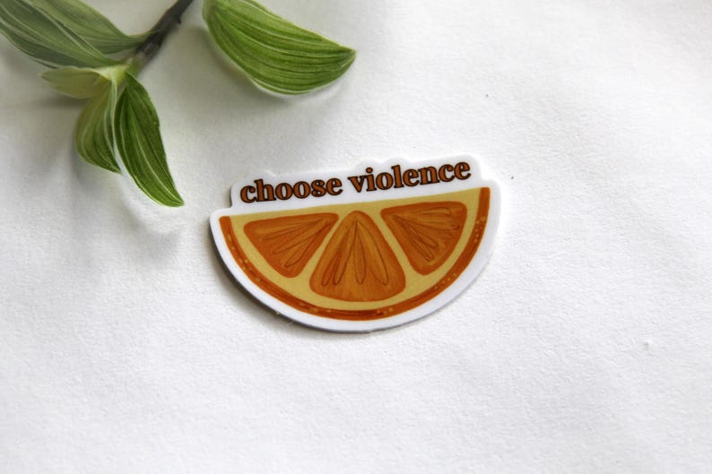 May include: A sticker featuring a slice of orange with the words "choose violence" in brown text. The orange slice is orange and brown with a white border. Green leaves are in the background.