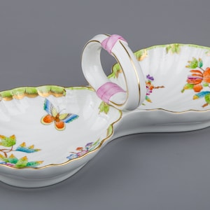 May include: A white porcelain serving dish with two sections, each shaped like a scallop shell. The dish has a pink ribbon handle and is decorated with green leaves, flowers, and butterflies.