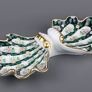 May include: A white porcelain serving dish shaped like two seashells with a black and green floral pattern. The dish has a gold trim and is perfect for serving snacks or appetizers.