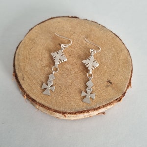 May include: A pair of silver dangle earrings with a geometric design. The earrings feature a square-shaped pendant with a cross-shaped pendant hanging below it.