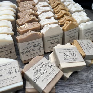 May include: An assortment of handmade soap bars with various scents and ingredients, including oatmeal milk, goat milk, and aloe vera. Each bar is wrapped with a label that includes the name of the soap and the ingredients. The soaps are arranged on a dark surface.