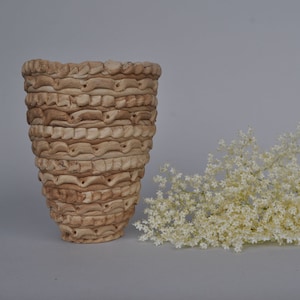 Op de afbeelding: Een lichtbruine keramische vaas met een getextureerd, golvend ontwerp. De vaas staat op een lichtgrijze ondergrond. Een tros witte vlierbloesems bevindt zich op de voorgrond.