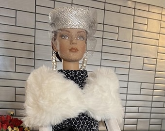 Può includere: Una bambola che indossa un abito bianco e nero con uno scialle di pelliccia bianca, un cappello argentato con velo, guanti bianchi e un braccialetto di perle. La bambola tiene una borsa nera e sta in piedi su una piattaforma bianca. Un cane di peluche rosa e soffice è seduto sul pavimento sullo sfondo.