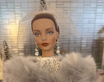 May include: A doll wearing a white sequined dress with a gray fur stole and a white veil. The doll has brown hair and is wearing pearl earrings.