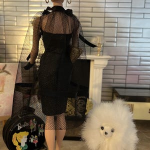 May include: A doll in a black dress with a sheer cape and gloves. The doll has a high bun hairstyle and is on a black stand. A white toy poodle and a black Barbie suitcase are also in the image.