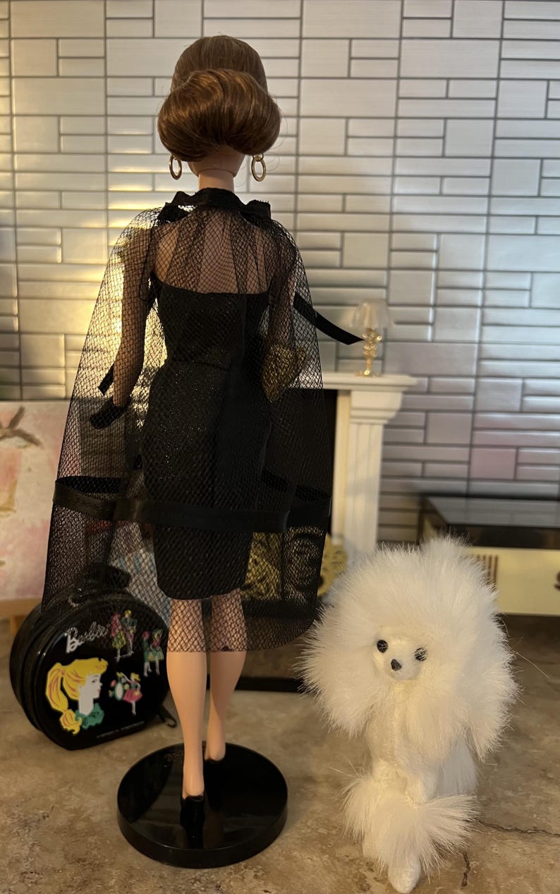 May include: A doll in a black dress with a sheer cape and gloves. The doll has a high bun hairstyle and is on a black stand. A white toy poodle and a black Barbie suitcase are also in the image.