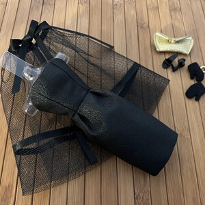 May include: A black doll dress with a bow, a black mesh veil, a gold clutch, black doll shoes, black doll gloves, and a gold-coloured charm. The items are arranged on a wooden surface.