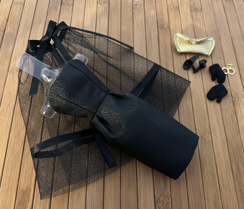 May include: A black doll dress with a bow, a black mesh veil, a gold clutch, black doll shoes, black doll gloves, and a gold-coloured charm. The items are arranged on a wooden surface.
