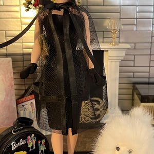 May include: A Barbie doll in a black mesh dress and cape, with matching gloves and heels. The doll has gold hoop earrings and a black bow. A black Barbie suitcase and a white fluffy dog are also in the image.
