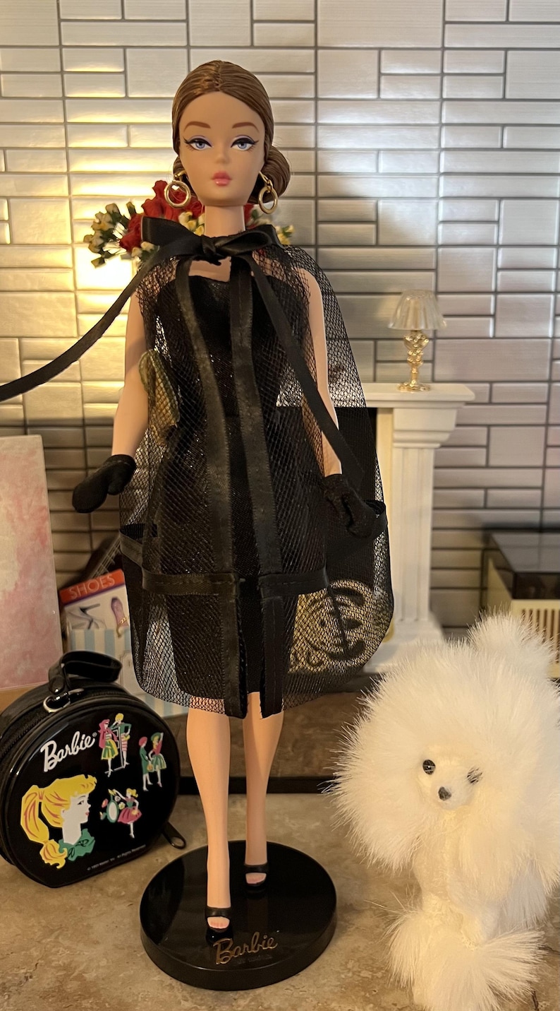 May include: A Barbie doll in a black mesh dress and cape, with matching gloves and heels. The doll has gold hoop earrings and a black bow. A black Barbie suitcase and a white fluffy dog are also in the image.
