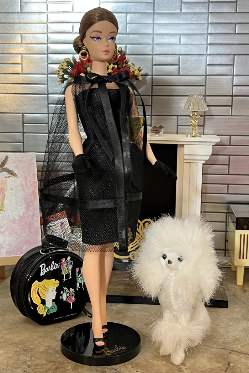 May include: A vintage Barbie doll in a black dress with a sheer cape, gloves, and heels. The doll has a floral headpiece and gold earrings. A black Barbie suitcase and a white fluffy dog are also in the image.