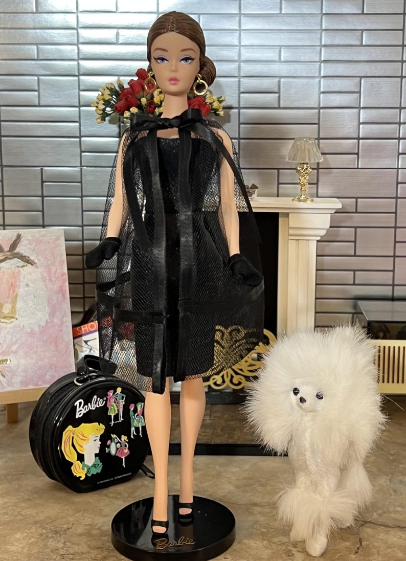 May include: A vintage Barbie doll in a black dress and cape, with a matching handbag and a white toy poodle. The doll is posed on a black stand. The background includes a fireplace and a painting. The doll's accessories include black gloves.