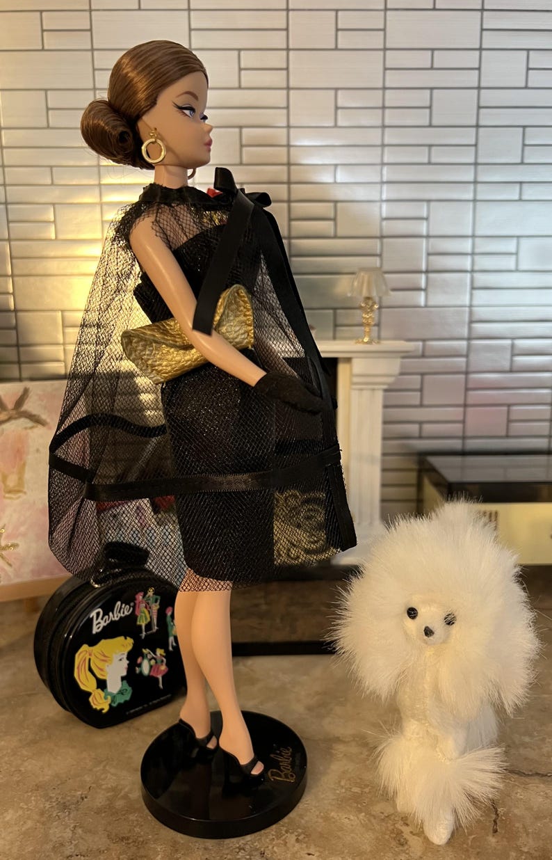 May include: A Barbie doll in a black dress with a sheer cape and gold clutch. The doll has a classic hairstyle and is standing on a black base. A small white dog is next to the doll, and a black Barbie suitcase is on the left.