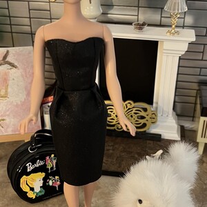 May include: A Barbie doll in a black strapless dress, black heels, and gold hoop earrings. The doll is standing on a black stand with the word "Barbie" on it. A black Barbie suitcase and a fluffy white dog are also in the image.