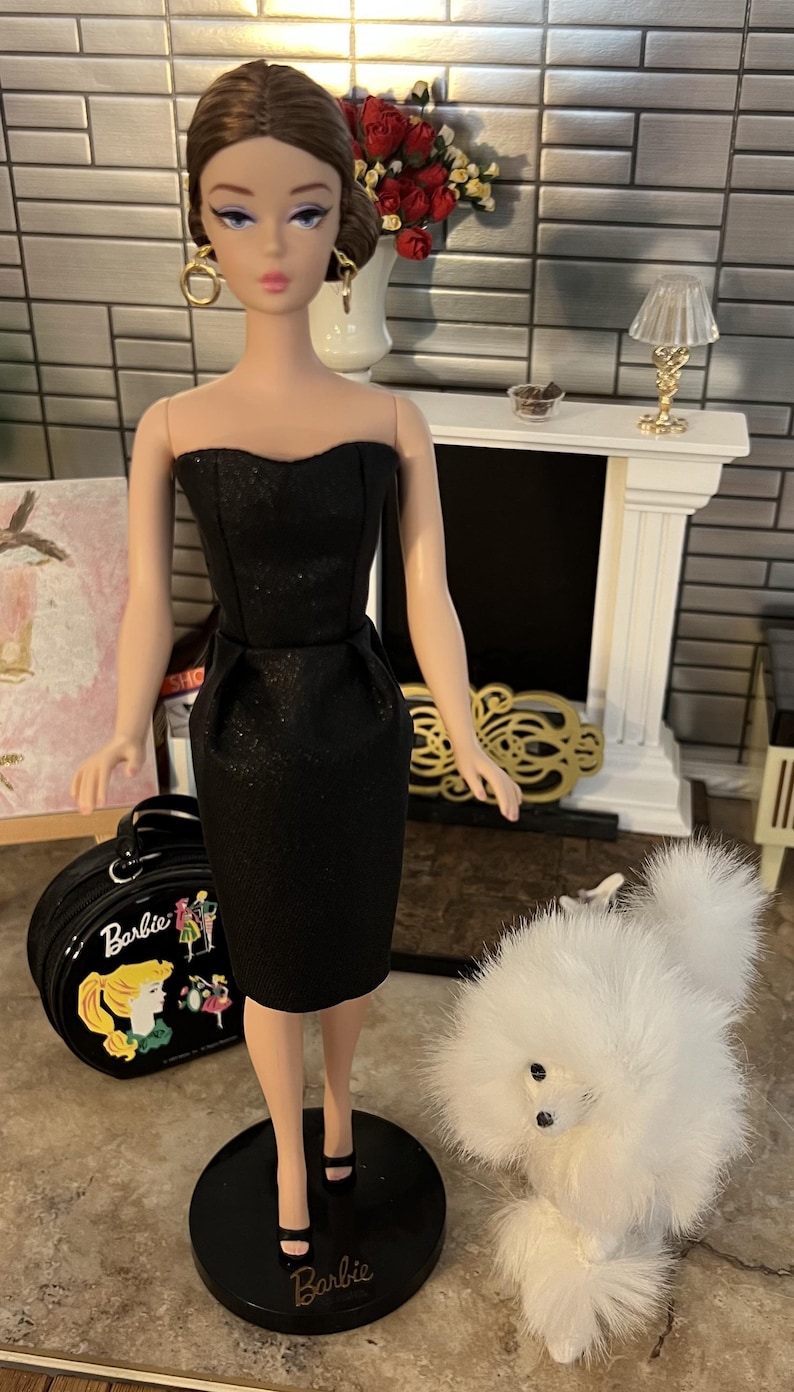 May include: A Barbie doll in a black strapless dress, black heels, and gold hoop earrings. The doll is standing on a black stand with the word "Barbie" on it. A black Barbie suitcase and a fluffy white dog are also in the image.