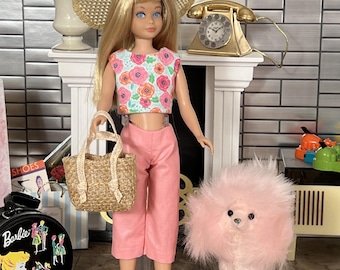 May include: A vintage Barbie doll in a floral crop top, pink trousers, and a straw hat, carrying a woven handbag. A pink poodle toy stands nearby. A gold telephone and a Barbie shoe case are in the background.