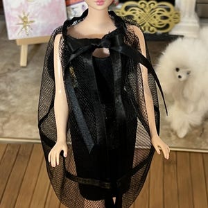 May include: A vintage Barbie doll in a black dress and a sheer black cape with a bow. The doll has brown hair and is on a black stand. A painting and a small white dog are in the background.