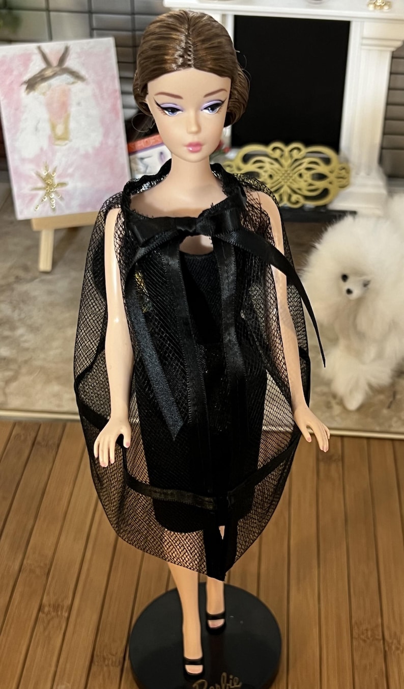 May include: A vintage Barbie doll in a black dress and a sheer black cape with a bow. The doll has brown hair and is on a black stand. A painting and a small white dog are in the background.
