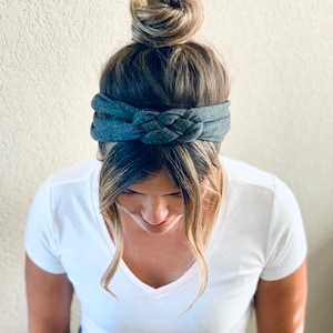 Op de afbeelding: Een donkergrijs gevlochten hoofdband gedragen door een persoon met blond haar in een knot. Het hoofdband is gemaakt van een zachte, rekbare stof.