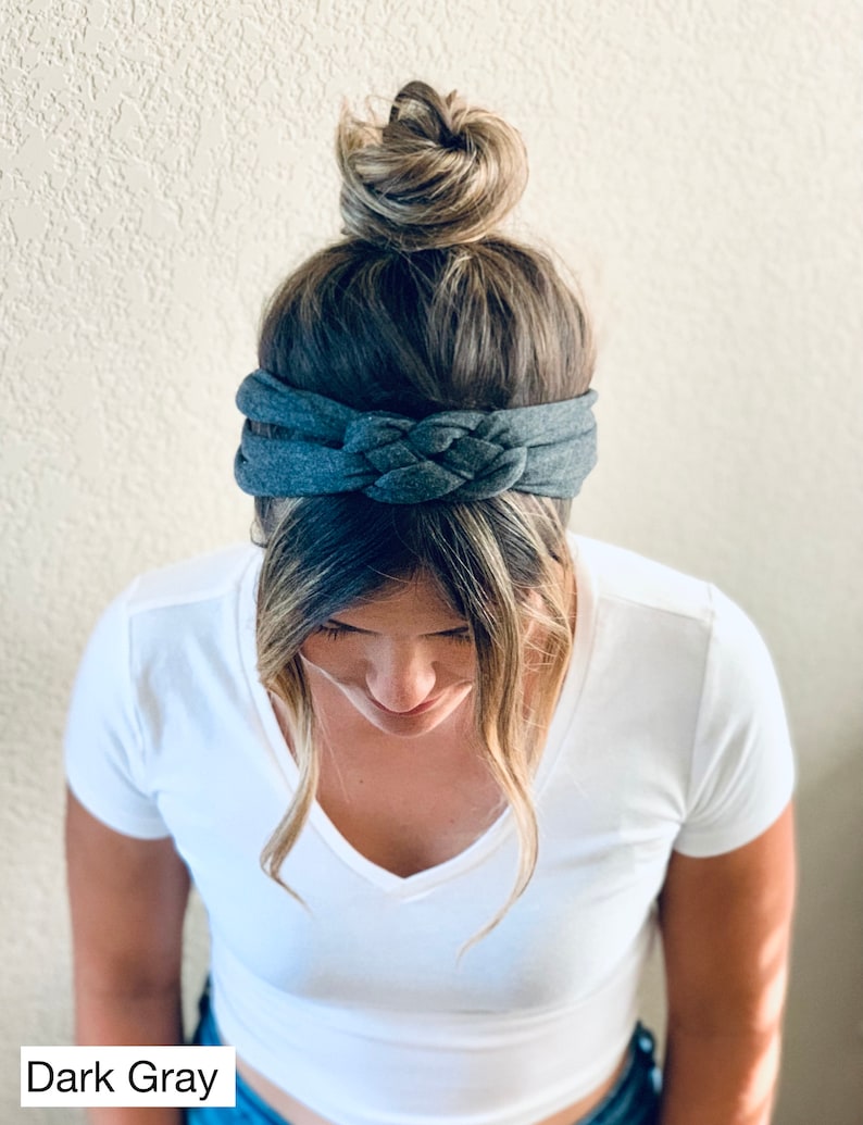 Op de afbeelding: Een donkergrijs gevlochten hoofdband gedragen door een persoon met blond haar in een knot. Het hoofdband is gemaakt van een zachte, rekbare stof.