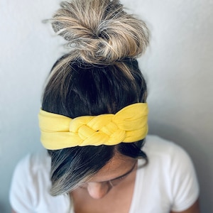 Op de afbeelding: Een gele geknoopte hoofdband van stof. De hoofdband wordt gedragen door een persoon met bruin haar dat in een knot is getrokken.