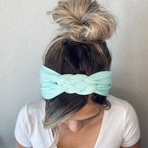 Op de afbeelding: Een turquoise geknoopt hoofdband van zachte stof. Het hoofdband wordt gedragen door een persoon met blond haar dat in een knot is geknoopt.