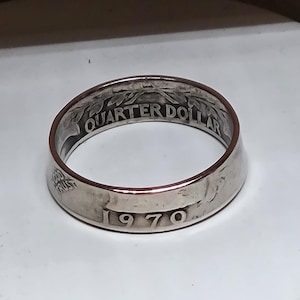 May include: A silver ring crafted from a 1970 quarter. The ring features the words "QUARTER DOLLAR" and "GOD TRUST" engraved on the band. The year "1970" is also visible on the ring's surface.