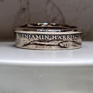 Könnte beinhalten: Ein silberner Ring mit der Inschrift "BENJAMIN HARRISON" um das Band. Der Ring hat ein detailliertes Design und scheint aus einer Münze gefertigt zu sein. Der Ring wird auf einer weißen Oberfläche präsentiert.