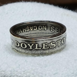 Puede incluir: Anillo plateado con las palabras "DOYLESTOWN" y "CASH VALUE" grabadas en la banda. El anillo, sobre un fondo blanco, resalta su brillo metálico y la inscripción detallada.