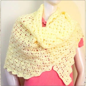 May include: A light yellow crocheted shawl with a scalloped edge. The shawl is draped over a mannequin, showcasing its openwork design. The yarn appears soft and the color is a pale, sunny yellow. The shawl is a triangle shape.