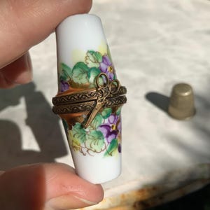 May include: A small, cylindrical porcelain trinket box with a white base, decorated with hand-painted purple flowers and green leaves. The box features a hinged, ornate brass band around the center, adorned with a small, attached brass scissor charm.