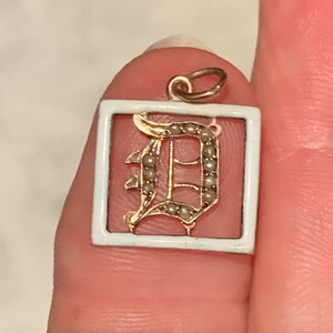 Op de afbeelding: Een gouden hanger met een wit emaille frame en een parel-ingelegde letter 'D' in een schrijflettertype.