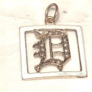 Op de afbeelding: Een gouden hanger met een wit ge&euml;mailleerd vierkant frame en een met parels ingelegde letter D.