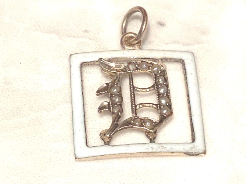 Op de afbeelding: Een gouden hanger met een wit ge&euml;mailleerd vierkant frame en een met parels ingelegde letter D.