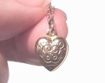 May include: A gold heart-shaped locket necklace with a delicate chain. The locket has a detailed engraved design.