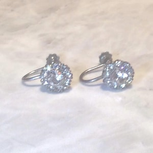 May include: A pair of silver-toned screw-back earrings, each featuring a central clear round stone surrounded by a halo of smaller clear stones. The metal components have a polished finish, and the design includes a curved wire mechanism.