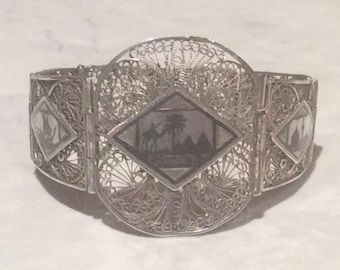 May include: A silver filigree bracelet with a central diamond-shaped panel featuring a silhouette of a camel and palm trees. The bracelet is decorated with intricate floral patterns and has a hinged design.