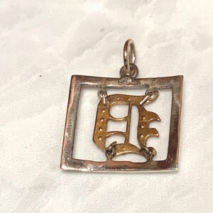 Op de afbeelding: Zilveren hanger met een gouden letter D in een vierkant frame.