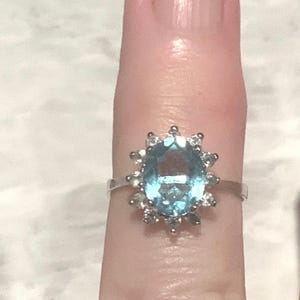 Op de afbeelding: Een zilveren ring met een grote ovale blauwe edelsteen omringd door kleinere heldere edelstenen.