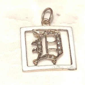 Op de afbeelding: Een gouden bedel met een wit ge&euml;mailleerd vierkant frame en een met parels bezette letter 'D' erin.