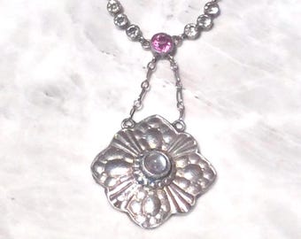 Puede incluir: Un collar de plata con un colgante en forma de flor. El colgante tiene una piedra transparente en el centro y está rodeado de piedras más pequeñas. El collar está adornado con pequeñas piedras rosas.