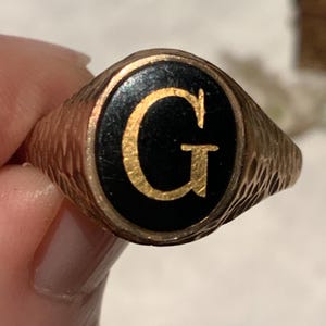 Può includere: Un anello con sigillo dorato con una faccia ovale nera. La faccia presenta la lettera "G" dorata. L'anello ha una fascia strutturata.