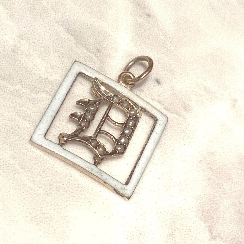 Op de afbeelding: Een gouden hanger met een wit ge&euml;mailleerd vierkant frame en een met parels bezette letter 'D' erin.
