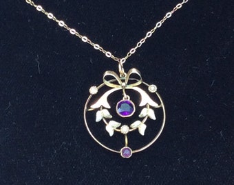 Puede incluir: Un collar de oro con un colgante que presenta un círculo con una piedra preciosa púrpura y un lazo en la parte superior. El círculo está decorado con pequeñas perlas blancas y hojas.