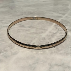 May include: A gold-coloured bangle bracelet with a textured surface. The bracelet has a simple, circular design and a clasp closure. The surface has a repeating pattern of small, rectangular indentations.