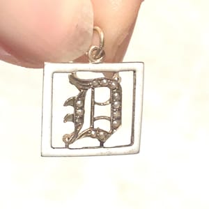 Op de afbeelding: Een zilveren bedel met een wit ge&euml;mailleerd vierkant frame en een gouden letter 'D' omrand met parels.