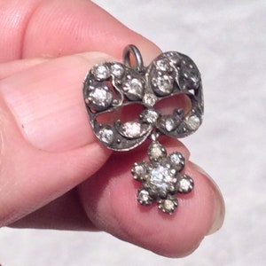 May include: A silver pendant with a delicate, ornate design featuring a heart-shaped frame with a flower-shaped drop. The pendant is adorned with numerous small, clear stones.